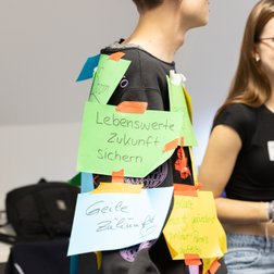 SustaiNable Conference 2024. Studierender wird mit Zetteln mit Aspekten zum Thema Nachhaltigkeit beklebt.