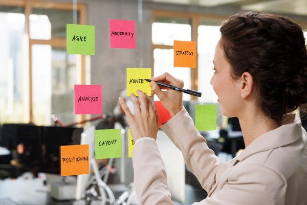 Junge Frau steht im Büro an einer Glaswand und beschriftet Post-its mit Staedtler Stiften