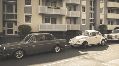 Alte Autos, u.a. ein Käfer parken vor einem Mehrfamilienhaus. 