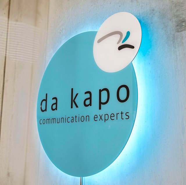 Das türkisfarbene da kapo Logo als 3D-Schild hängt LED-beleuchtet an der Betonwand in den Büroräumen
