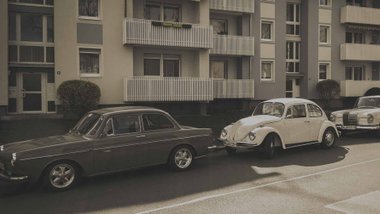Autos u.a. ein Käfer parken am Straßenrand vor einem Mehrfamilienhaus. 