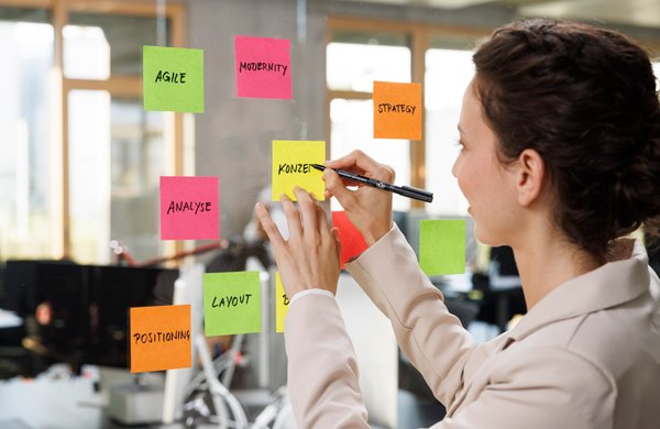 Junge Frau steht im Büro an einer Glaswand und beschriftet Post-its mit Staedtler Stiften