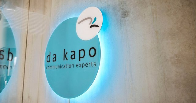 Das türkisfarbene da kapo Logo als 3D-Schild hängt LED-beleuchtet an der Betonwand in den Büroräumen