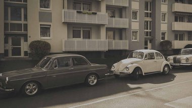 Autos u.a. ein Käfer parken am Straßenrand vor einem Mehrfamilienhaus. 