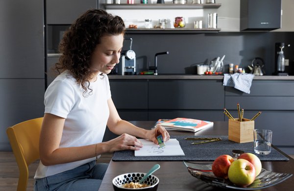 Junge Frau sitzt am Küchentisch und malt mit Staedtler Stiften